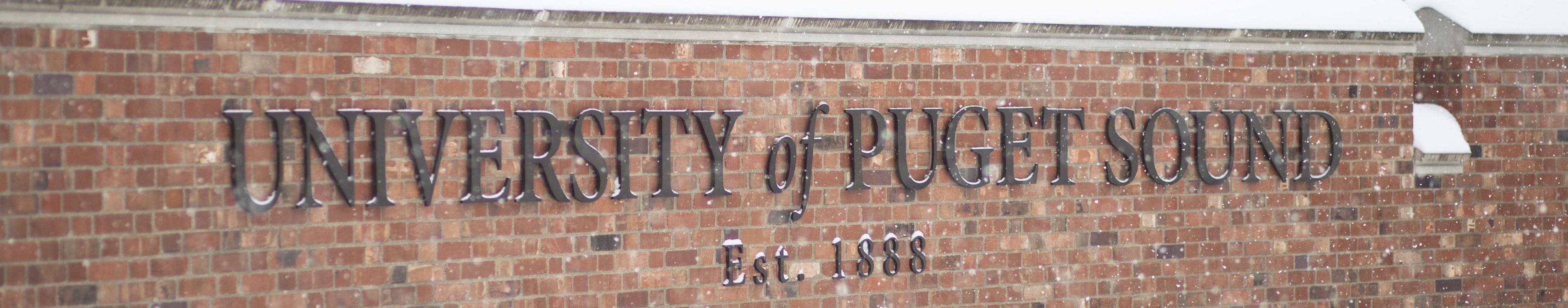 A brick University of Puget Sound sign sits covered in snow. 
