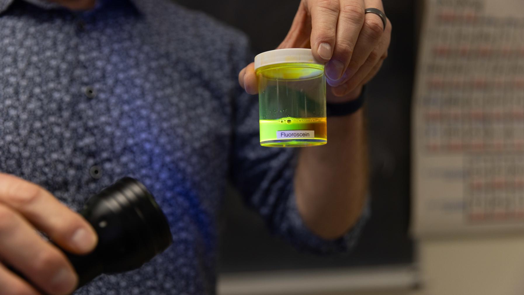 Prof. Dan Burgard shines an ultraviolet light on a liquid in a glass container to produce a green/orange.