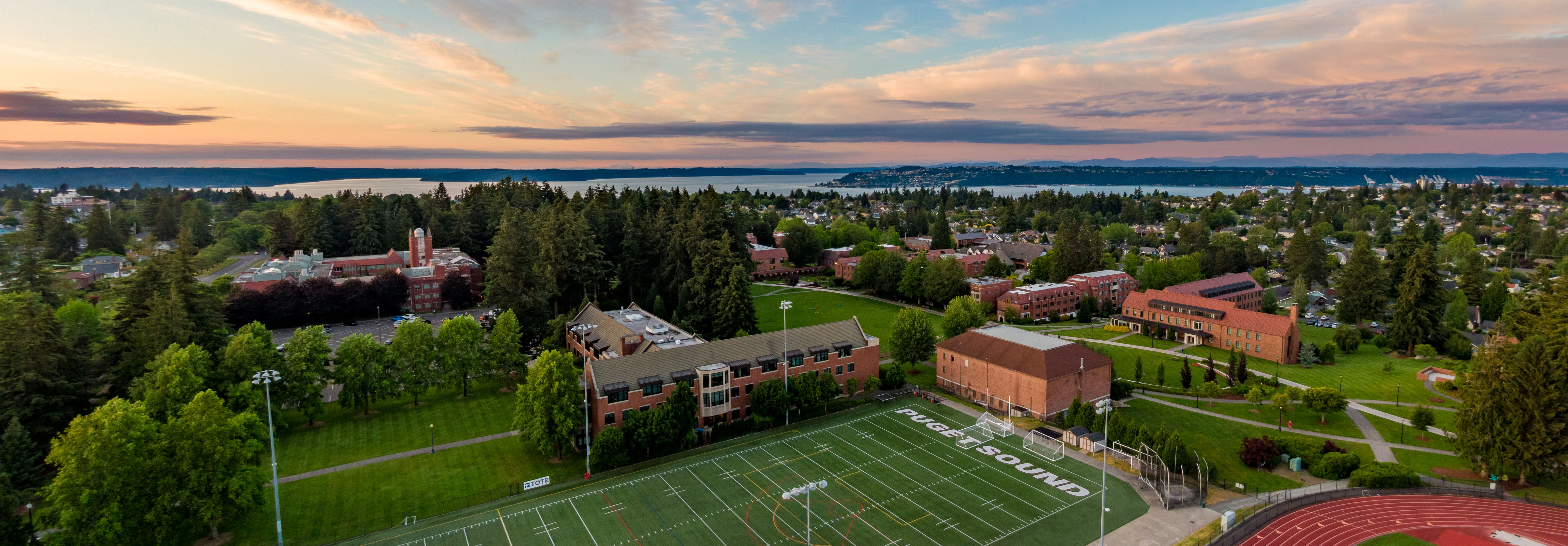 Aerial view of the campus as the sun sets, with red brick buildings and trees, and the Puget Sound in the background.