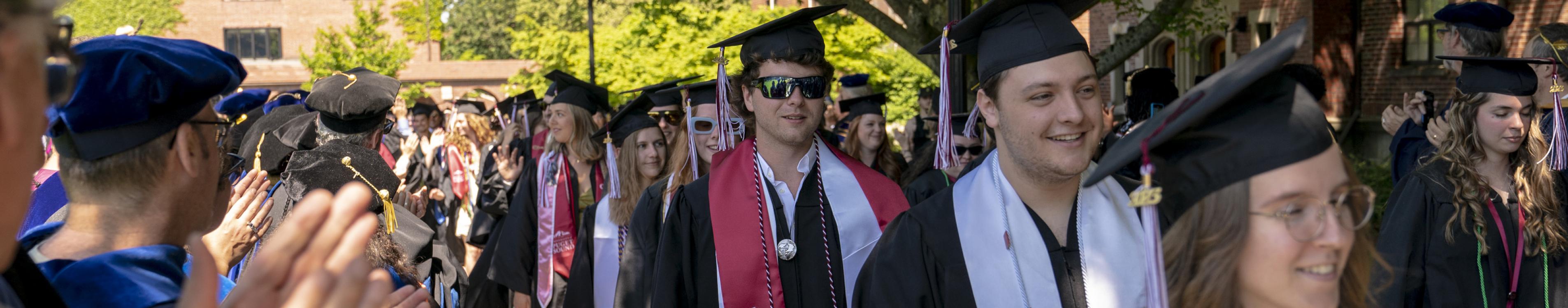A line of students in graduation regalia walk toward the camera. 