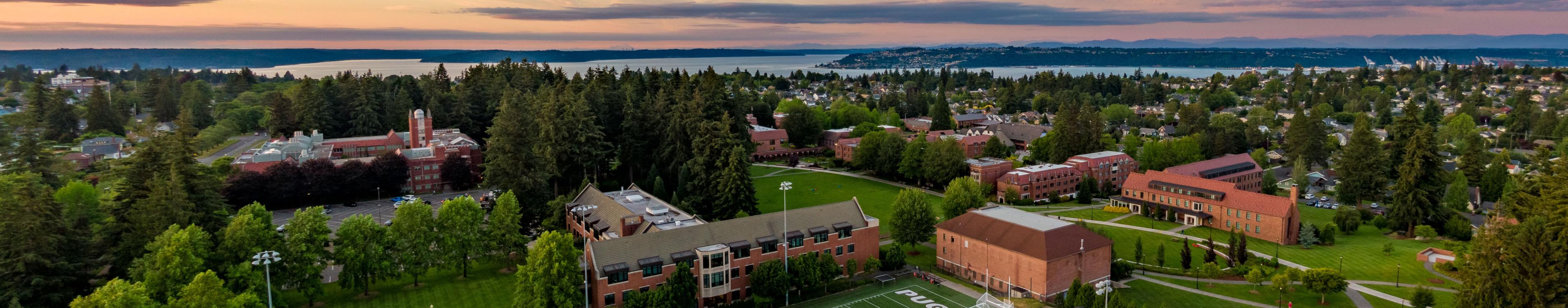 Aerial view of the campus as the sun sets, with red brick buildings and trees, and the Puget Sound in the background.