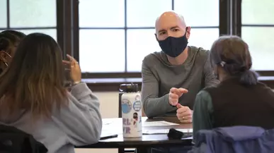 A masked male teacher speaks to students with windows behind him