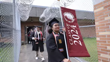 Inmate walks down gated walkway carrying a Class of 2024 banner while wearing graduation regalia. 