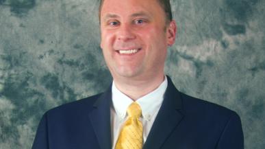 Director of Institutional Research Kevin Riordan smiles at the camera in a blue suit and yellow tie. 