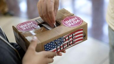 A hand drops a ballot into a brown box with the words VOTE HERE written on it.