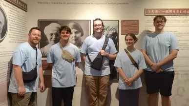 Five students in blue shirts stand in front of a wall with the Trimble family history on it.