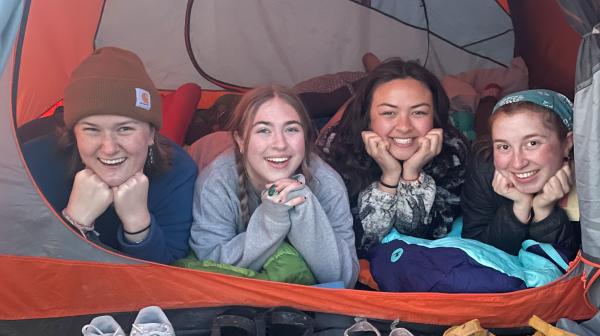 Students lay on their stomachs with their heads propped up on their elbows smiling out the open tent door.