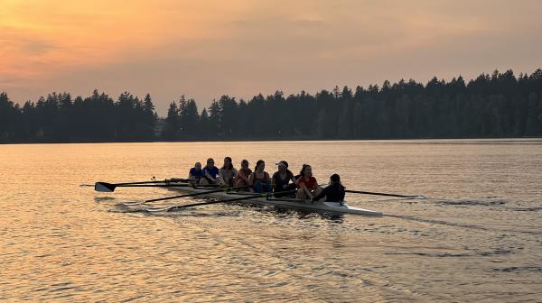 A crew boat filled with rowers row in a sunrise-lit, tree-lined lake.