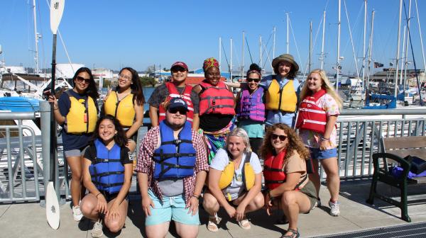 A group of students in life vests stand on a harbor dock and smile at the camera.