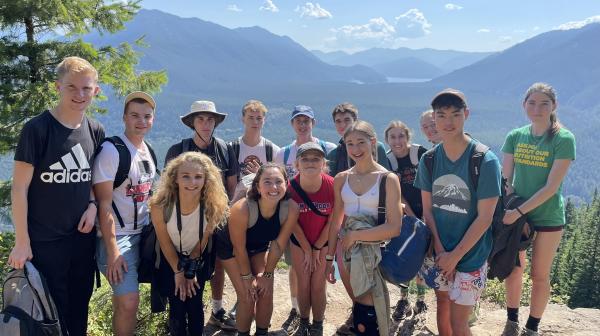 A group of students stand on a sandy ledge overlooking a tree covered valley.
