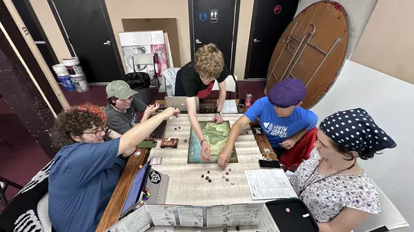 Students sit and stand around a large table spread with papers, a map, mini figures, and other game supplies.