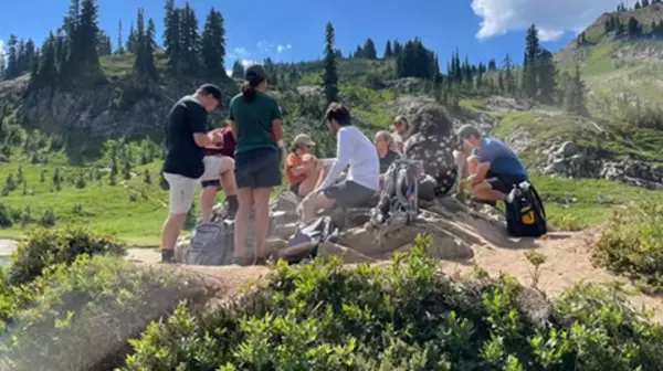 Students gather and sit and stand on a sandy broad trail in a sun covered hill, writing in notebooks.