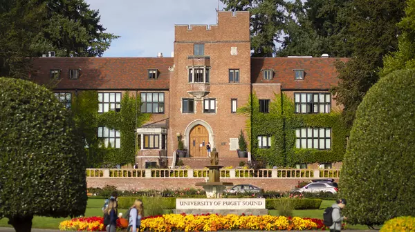View of a red brick building with ivy growing on it and trees on either side, and a University of Puget Sound sign in the foreground.