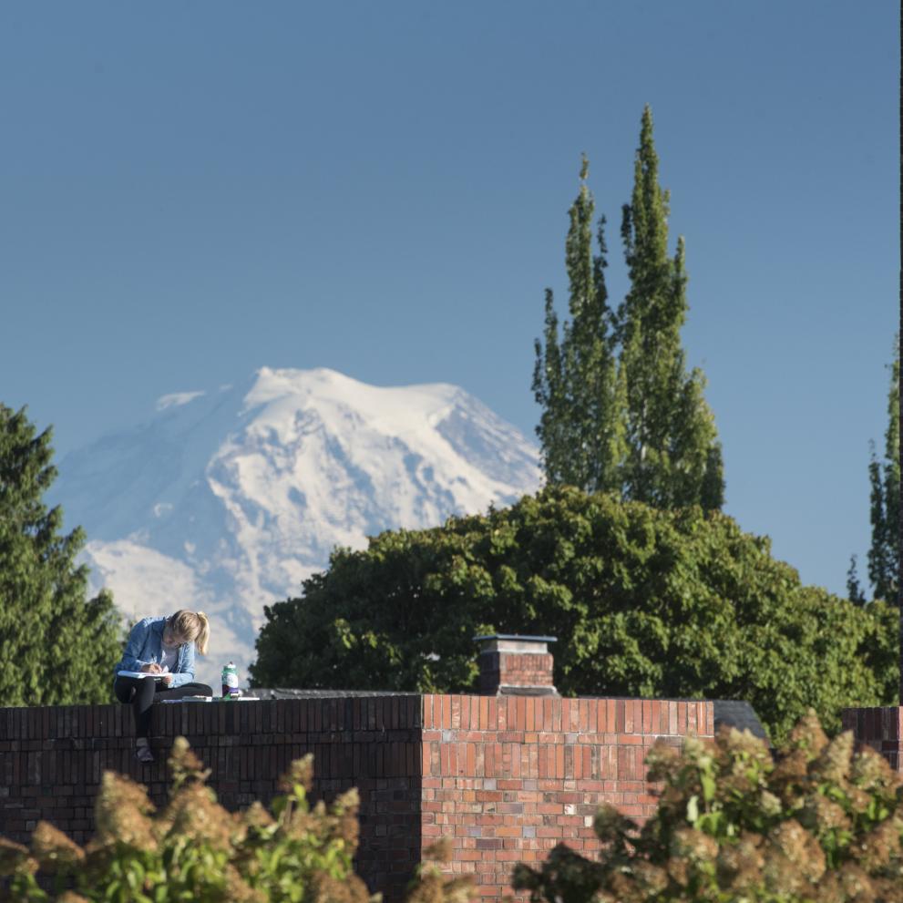 A student sits atop a wall enclosing an outdoor space near Weyerhaeuser Hall with Mount Rainier perched behind.