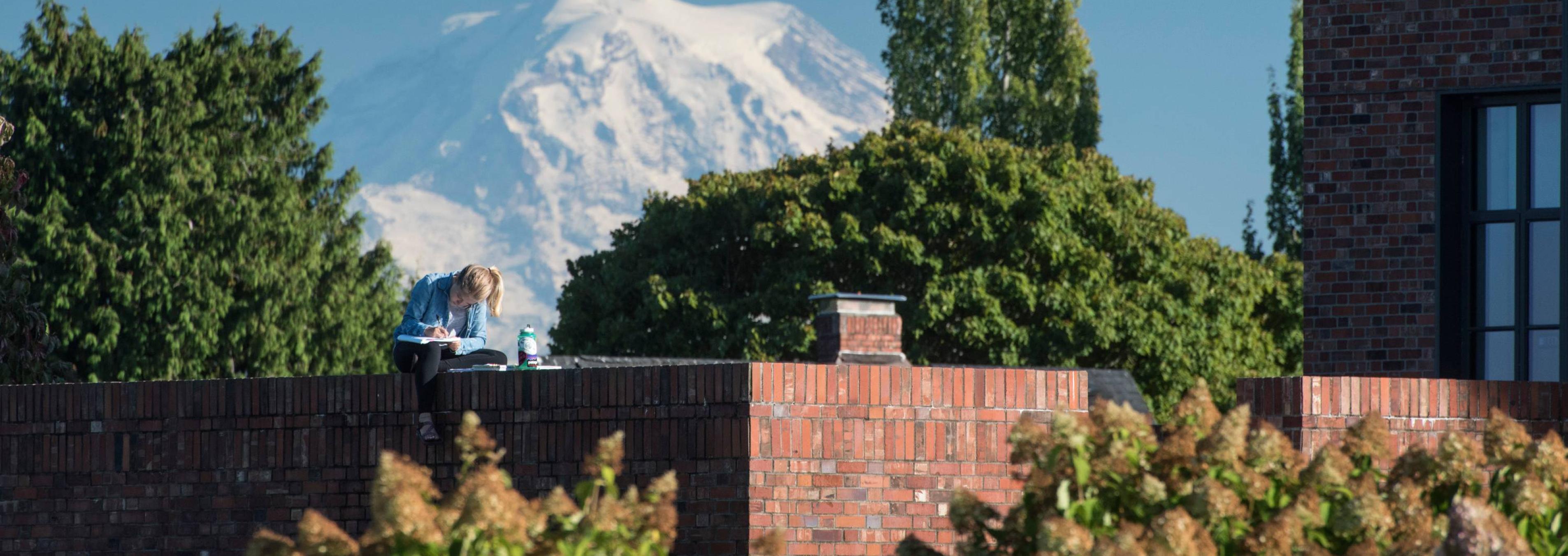 A student sits atop a wall enclosing an outdoor space near Weyerhaeuser Hall with Mount Rainier perched behind.