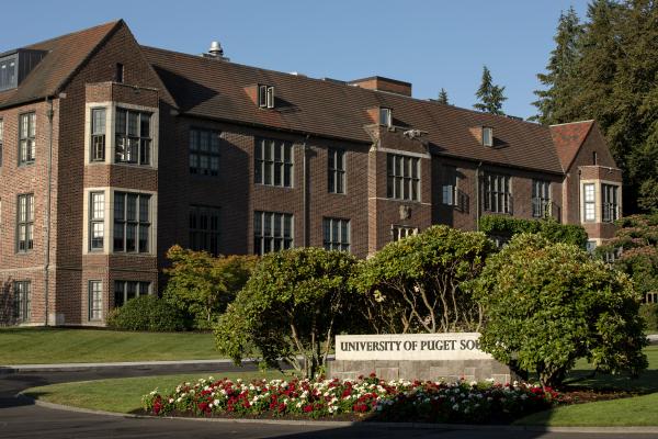 The University of Puget Sound sign sits behind flowers in front of a brick building. 