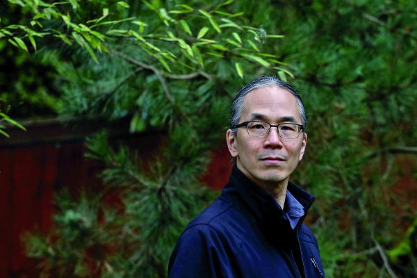 Ted Chiang looks over his shoulder at the camera. He is wearing glasses and a blue coat. 
