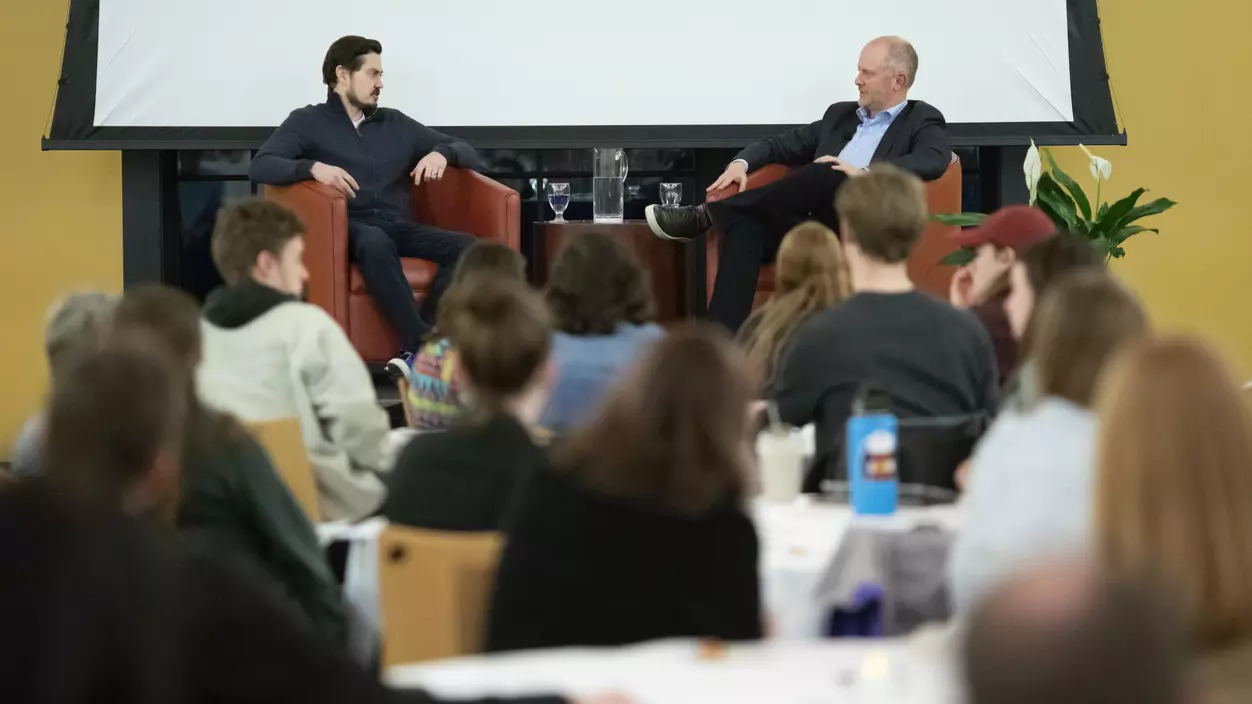 Two men sitting on a stage with an audience of people in front of them