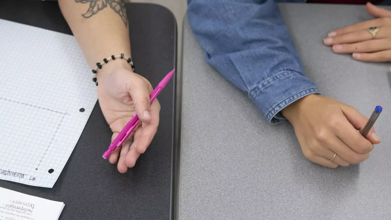 Two people's arms, one with tattoos, holding pens and writing at a table