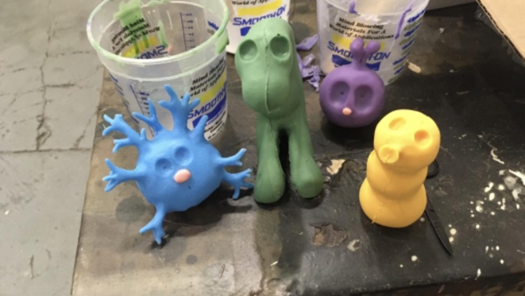 Blue, green, purple, and yellow clay figures in various stages of progress stand on the corner of a work table