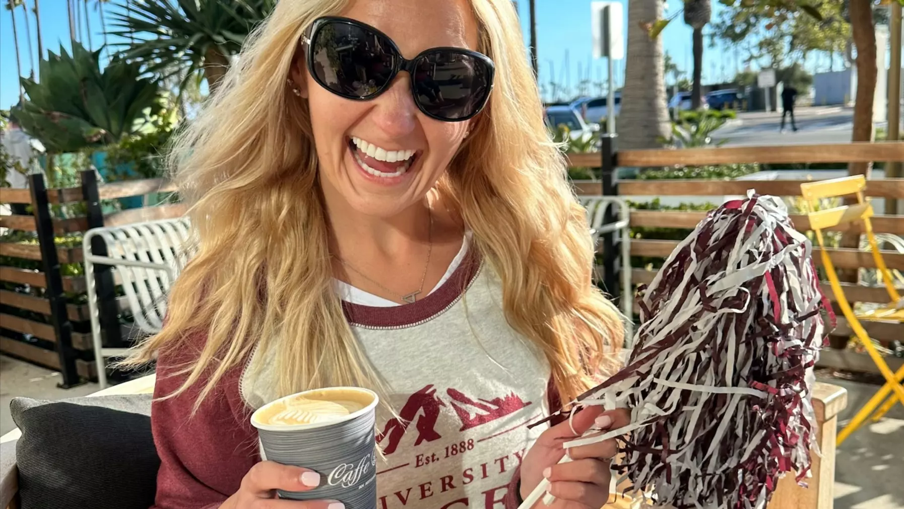 A seated, smiling alumni Logger holding a coffee and maroon and white pompom