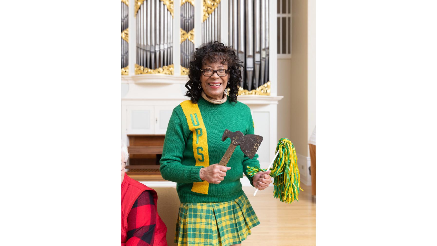 An alumna from the Class of 1973 dressed in a green and gold cheerleading uniform and holding the hatchet at Summer Reunion Weekend 2023.