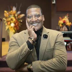 Man smiles at camera in a gold/tan suit with a black button up shirt underneath. 