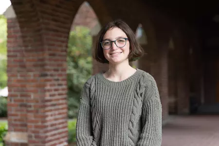 Daisy Davidson-Innis stands under brick arches wearing a green shirt and glasses. 