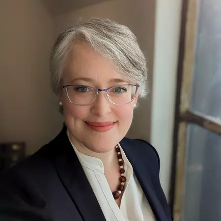 Woman with short silver hair and glasses smiling in front of a window.